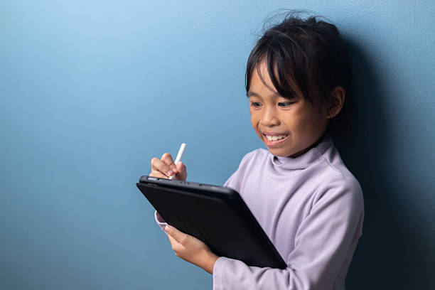 istock Smiling Asian child using digital tablet with stylus for online learning on blue wall background, concept of happy education, creative learning, and digital technology for kids. 2242792480