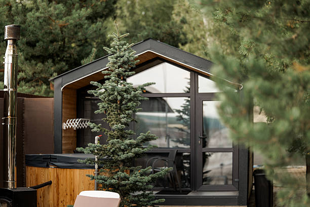 A modern cabin with large windows is surrounded by lush greenery, creating a peaceful and inviting atmosphere. The cabin features a unique architectural design.