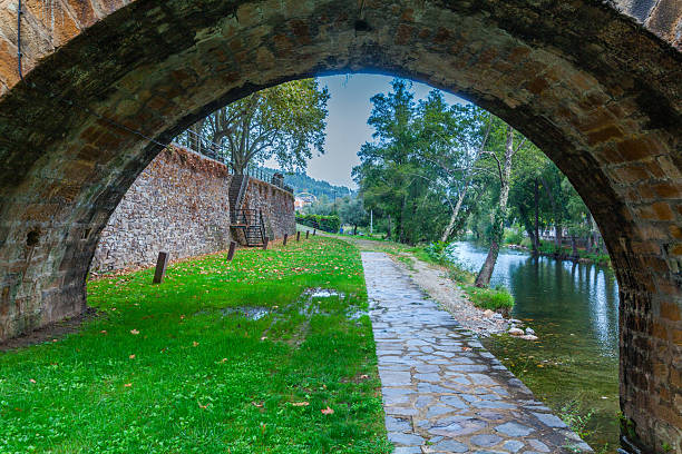 Viewed from under a historic stone arch, a cobblestone path winds beside a calm river through a lush green park, framed by trees under an overcast sky