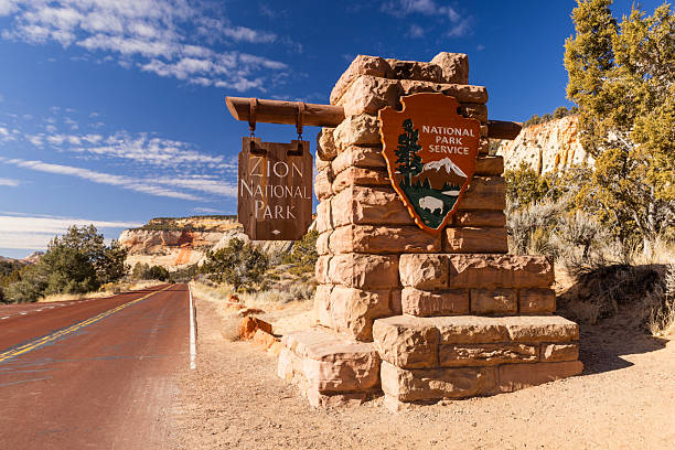 The Zion National Park sign on a beautiful day in Utah with white fluffy clouds and blue skies creating a welcoming feel to the park.