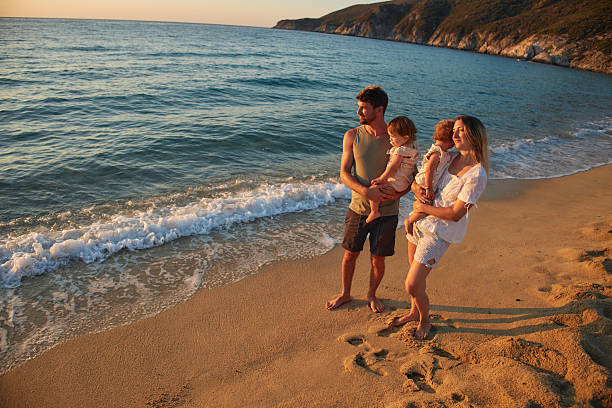 Parents holding toddlers standing on a beach watching the ocean waves at sunset