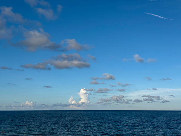 A tranquil view of the Atlantic Ocean as scattered cumulus clouds drift across a bright blue Florida sky. The deep blue water meets the horizon seamlessly, with small groups of swimmers near the shore. This minimalist seascape captures the calm, open expanse of Fort Lauderdale’s coastline under soft afternoon light.