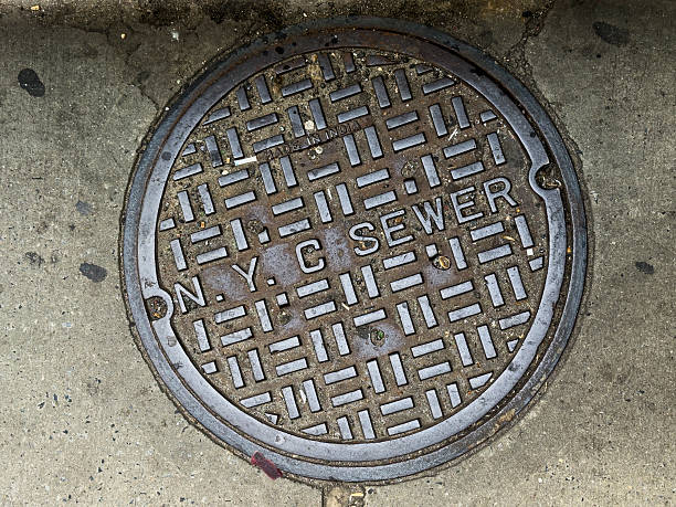A close-up view of a classic New York City sewer cover embedded in the concrete sidewalk. The bold, utilitarian typography and geometric grid pattern reflect the enduring industrial character of the city’s infrastructure—an everyday detail that embodies the rhythm and texture of urban life.