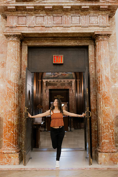 Woman smiling as she opens a large exit door in a grand marble-columned hall, stepping toward light and new possibilities in nyc