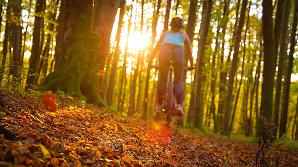 LENS FLARE, LOW ANGLE VIEW: Fallen autumn leaves fly in the air as female mountain biker rides along forest path. Fun and adrenaline filled outdoor sports activity in colourful woods in fall season.