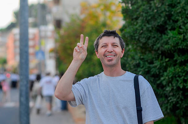 A young man beams with a radiant smile as he holds up one finger, embodying a message of unity, focus, or singularity. His gesture suggests a sense of importance or perhaps a reminder to focus on one key thing. Clad in a denim shirt, his casual style adds an approachable warmth to the image. The neutral backdrop keeps the emphasis on his expression and gesture, creating an inviting and positive visual statement about simplicity and strength in singularity.