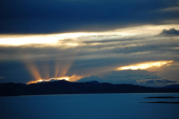 Sunbeams break through clouds, casting light over a calm lake with distant mountains under a dramatic sky.