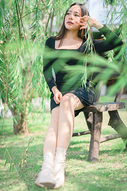 A young woman sits gracefully on a wooden bench, surrounded by lush greenery and soft sunlight, embodying elegance and tranquility in a beautiful outdoor setting.