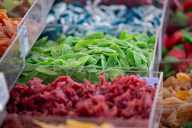 A vibrant assortment of dried fruit slices displayed in clear plastic containers at a market stall. The mix of green, red, and orange hues highlights the natural texture and variety of the fruits, creating an appealing and healthy visual composition.