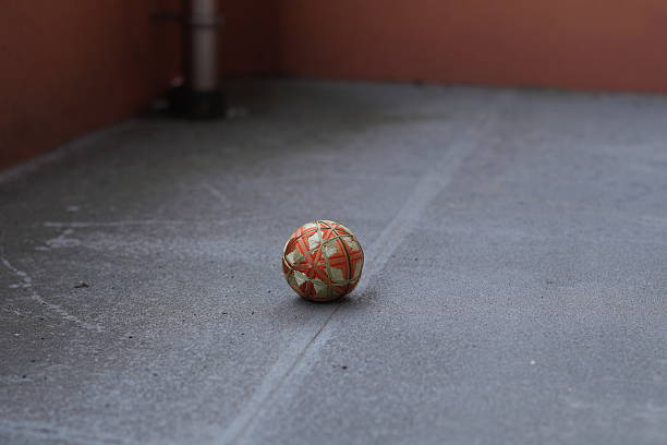 A small orange and green ball is sitting on a grey concrete floor. The ball is not in motion and he is resting. The scene is quiet and uneventful, with the ball being the only object in focus