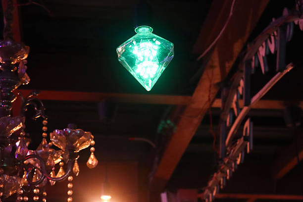 A captivating diamond-shaped glass lantern, glowing with a vivid green light, hangs in a dark, atmospheric room, complemented by the presence of an ornate chandelier.