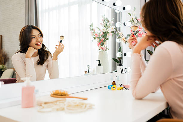 istock Smiling woman applying makeup at a bright vanity, looking into a large mirror with cosmetics, flowers, and accessories on the table in a sunlit modern bedroom. 2242929600