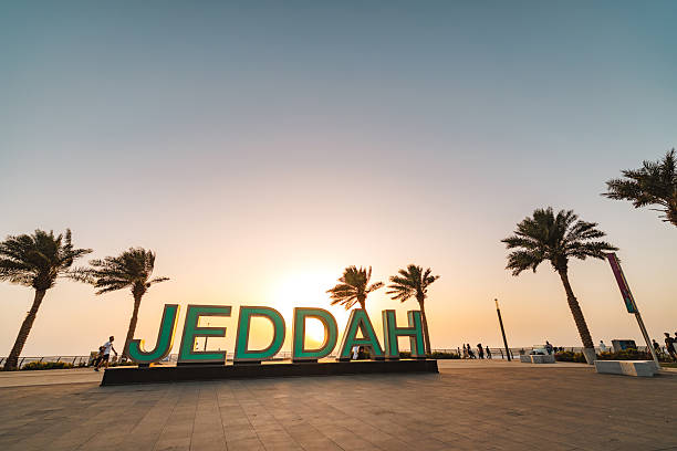 The iconic JEDDAH sign stands prominently on the Red Sea waterfront. Palm trees frame the scene as the sun sets, casting a warm glow over the area. People are seen walking nearby, enjoying the tranquil evening atmosphere.