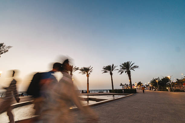 Blurry figures walking briskly along the palm-lined promenade at Jeddah Corniche during evening. The sky is transitioning from day to night, with streetlights illuminating the path. Palm trees and public sculptures add to the scenic backdrop.