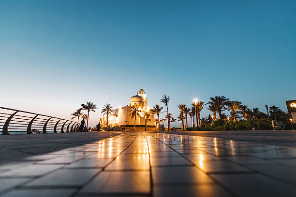 Wide shot capturing the illuminated Al-Rahma Mosque at Jeddah Corniche during dusk. The mosque is surrounded by palm trees and a paved walkway, with the sky transitioning from day to night. Soft lighting creates a tranquil atmosphere along the waterfront.