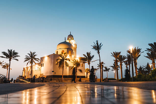 Al-Rahma Mosque illuminated against the evening sky at Jeddah Corniche. Palm trees line the walkway as people stroll by, creating a serene atmosphere. The reflective tiles on the ground add a warm glow to the scene.
