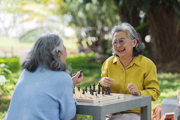 Two senior women enjoy a chess game outdoors surrounded by nature. One reacts with excitement while the other makes a move. A joyful scene of friendship, strategy, and mental activity.