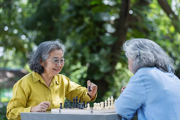 Two senior women enjoy a chess game outdoors surrounded by nature. One reacts with excitement while the other makes a move. A joyful scene of friendship, strategy, and mental activity.
