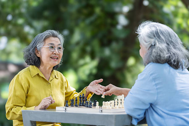 Two senior women play chess outdoors on a shiny metal table. One focuses on her move while the other watches attentively. A calm and thoughtful moment of friendship and mental exercise.