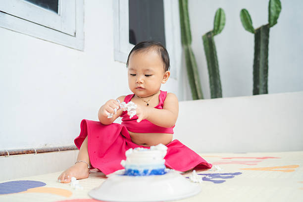 Baby girl holding frosting while exploring cake indoors, concept of sensory learning and milestone play