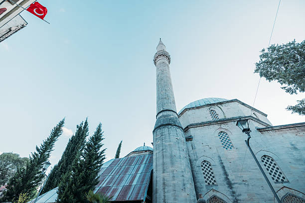 Ancient stone mosque with a tall minaret under clear sky and Turkish flag waving. Traditional Islamic architecture with domes and arched windows. Religious building in Turkey.
