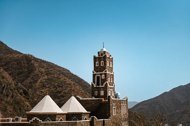 A tall, traditional stone tower stands prominently against a clear blue sky, surrounded by rugged mountain terrain. The architectural details of the structure are highlighted by the bright sunlight, showcasing its historical significance in the Rijal Almaa village.