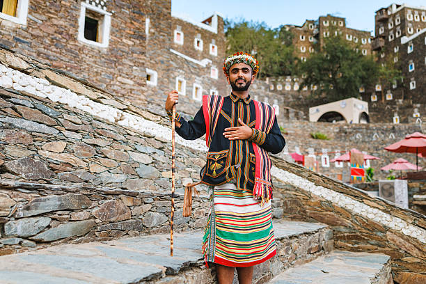 Man standing on stone steps wearing traditional Asiri dress with colorful patterns. Holding a decorative staff, he poses in front of the historic stone buildings of Rijal Almaa, conveying a sense of pride and cultural heritage.