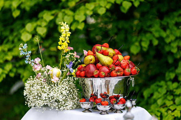 Fresh fruit, vegetables, and snacks are arranged on a table set up on a sandy beach, ready for a healthy vegan meal