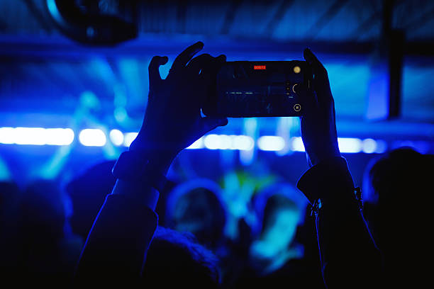 A person holding a smartphone while recording a live concert in a dark venue with blue stage lights