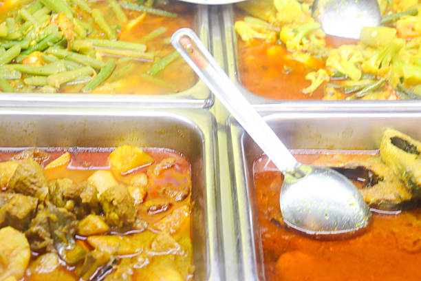 A close-up shot of a food buffet, featuring various colorful and aromatic curry dishes in metal trays, with a serving spoon ready to portion out a meal.