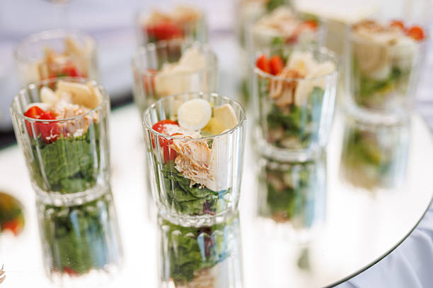 Multiple servings of salad in transparent glasses arranged on a reflective surface for outdoor catering or event dining.