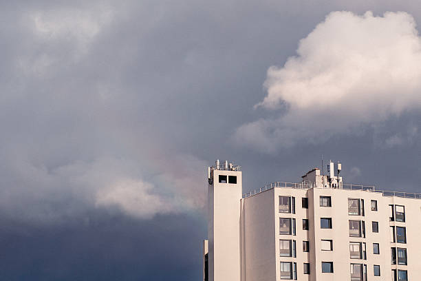 Minimal urban scene showing a white residential building under a stormy sky with a faint rainbow. Concept of hope, change, and weather.