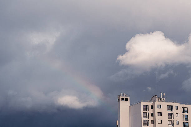 Minimal urban scene showing a white residential building under a stormy sky with a faint rainbow. Concept of hope, change, and weather.