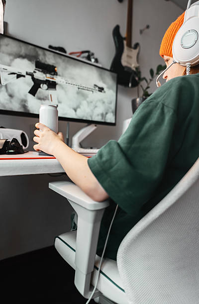 istock Young gamer sits in ergonomic chair, wearing headset, and reaching for energy drink while gaming. Modern gaming setup with sleek desk, large monitor, mechanical keyboard, high-performance accessories. 2242995120