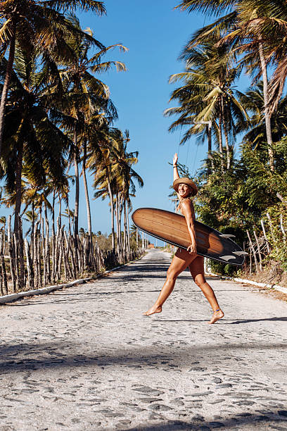istock Young woman walking barefoot along a tropical road with a surfboard 2243000949