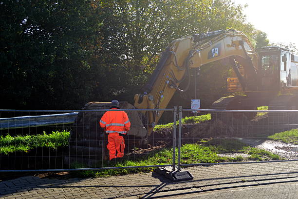 Leuven, Flemish-Brabant, Belgium - October 21, 2025: A road worker in orange reflective workwear stands in a ditch, overseeing the excavation of earth with a Cat excavator