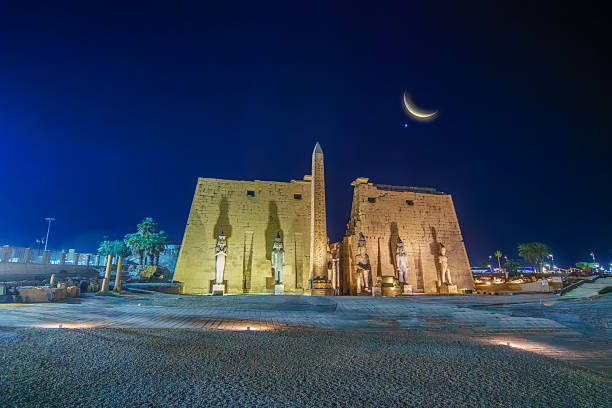 Wide angle establishing shot of the illuminated Luxor Temple Pylon statues and obelisk at night. A stunning crescent moon and the planet Venus are visible in the dark blue sky. Shot in 2025.