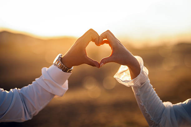 A close-up of two hands forming a heart shape against a beautiful sunset background. This romantic moment symbolizes love, connection, and togetherness. Perfect for themes related to relationships, weddings, and emotional connections