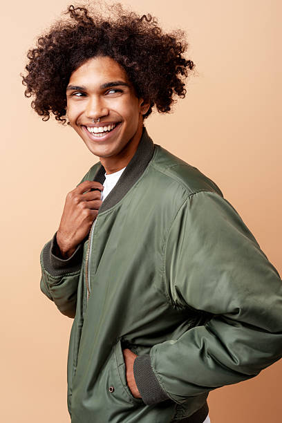 Young Afrolatino man with curly hair smiling and looking away, standing in a green bomber jacket on a plain studio background