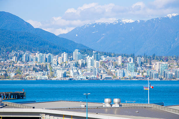 The serene beauty of West Vancouver is vividly captured from across the water, showcasing the gentle gradient of snow-dusted peaks descending into the lush coniferous forests. The quiet waters mirror the blue sky, punctuated by soft, drifting clouds, creating a harmonious blend of nature's elements in one tranquil vista.