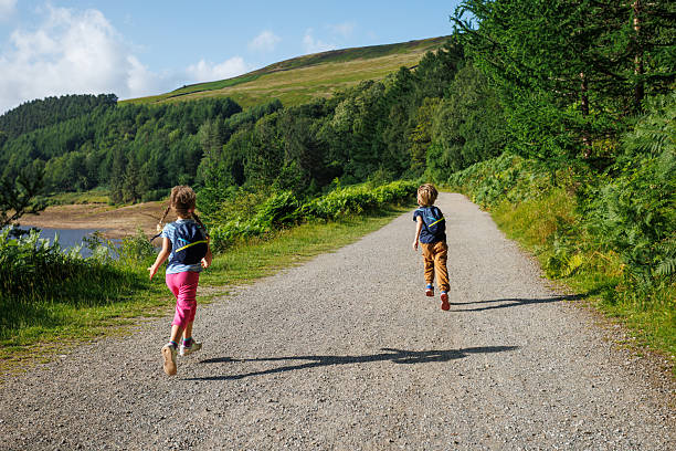 In the midst of a sunny woodland, two kids hikers, with packs are racing along a pebble-strewn path in Upper Derwent Valley, England