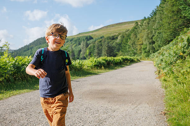 Smiling, a boy in glasses enjoys a sunny day as he walks down a rural road lined with plants