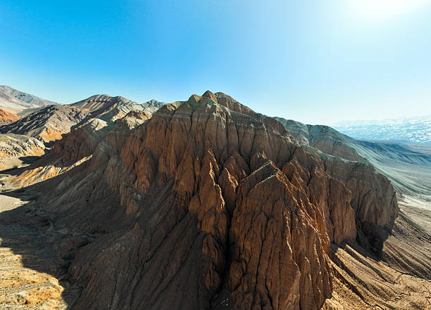Close-up aerial wide shot of a reddish-brown, heavily textured mountain peak featuring jagged vertical erosions under a stark, clear blue sky