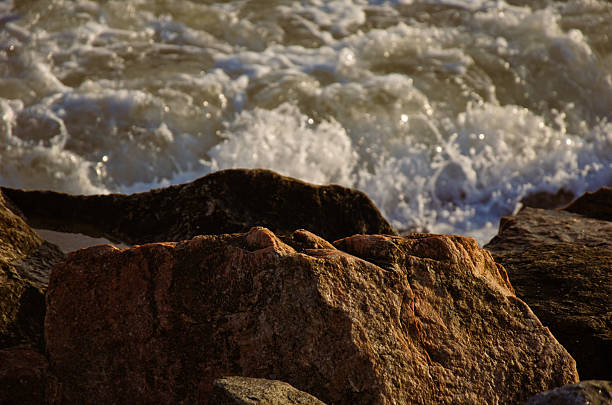 Close-up of textured coastal boulders bathed in warm, dramatic sunlight. Turbulent ocean waves crash behind the rocks, creating powerful white foam and spray. Captures the rugged beauty and raw power of nature.