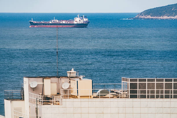 Large oil tanker ship cruising across deep blue sea on a sunny day, captured from a high vantage point behind a modern rooftop with satellite dishes and HVAC systems, with distant green headland