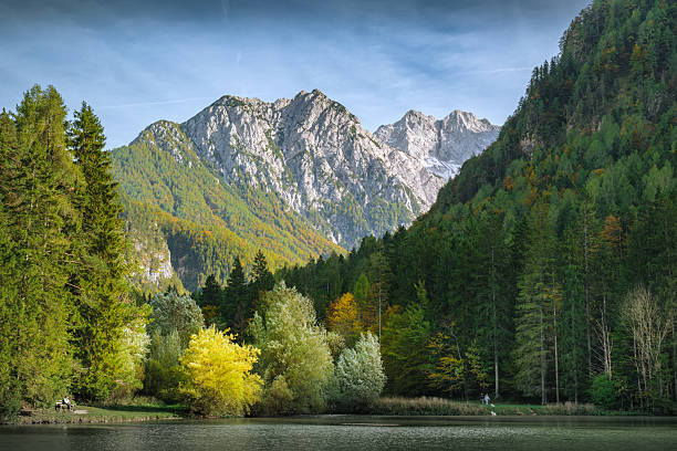 A lone person sits on a bench by a lake, enjoying the view of the colorful autumn foliage and majestic mountains. In the distance, two people walk their dogs along the shore, appreciating the serene natural landscape.