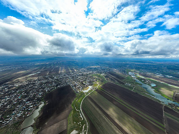 Expansive, high aerial perspective of villages and a distant urban area nestled within a vast, striped plain under a huge sky filled with bright, dramatic clouds