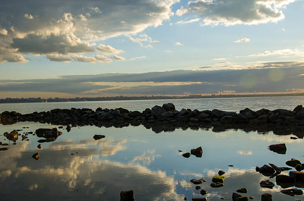 A tranquil scene during the golden hour, showing dramatic clouds reflecting perfectly in calm water amidst dark shoreline rocks. The horizon reveals a faint silhouette of a distant city or industrial area under a wide, moody sky.