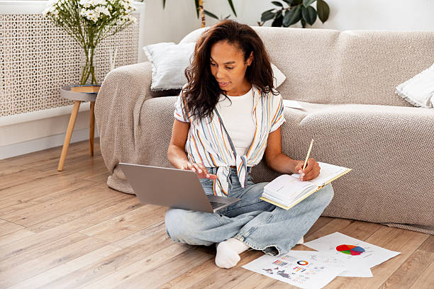 istock Smiling happy African American woman using laptop taking notes planning project sitting on floor 2243091189
