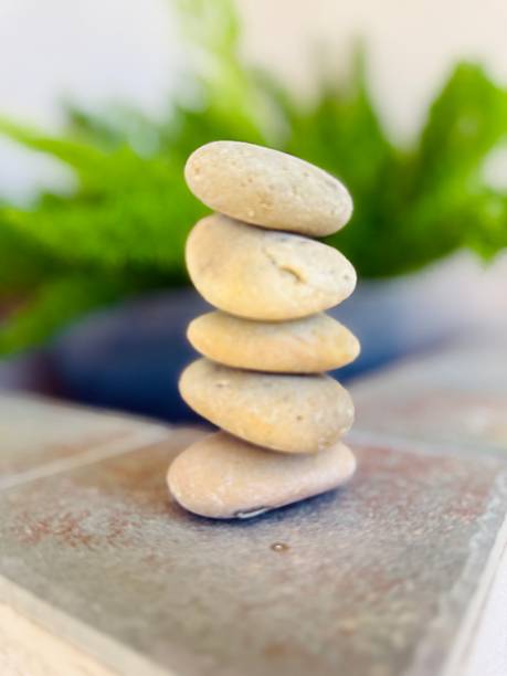 A stack of smooth stones arranged on a wooden deck, casting a shadow in natural light. Surrounding river stones add texture, evoking balance, tranquility, and nature's elegance.